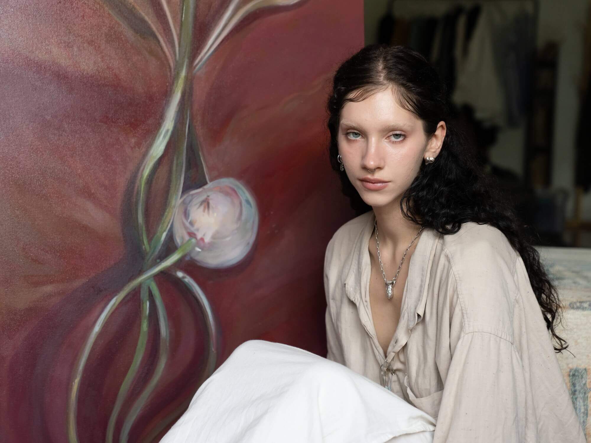 Woman sitting next to abstract painting, wearing a neutral-colored outfit, in an artist's studio.