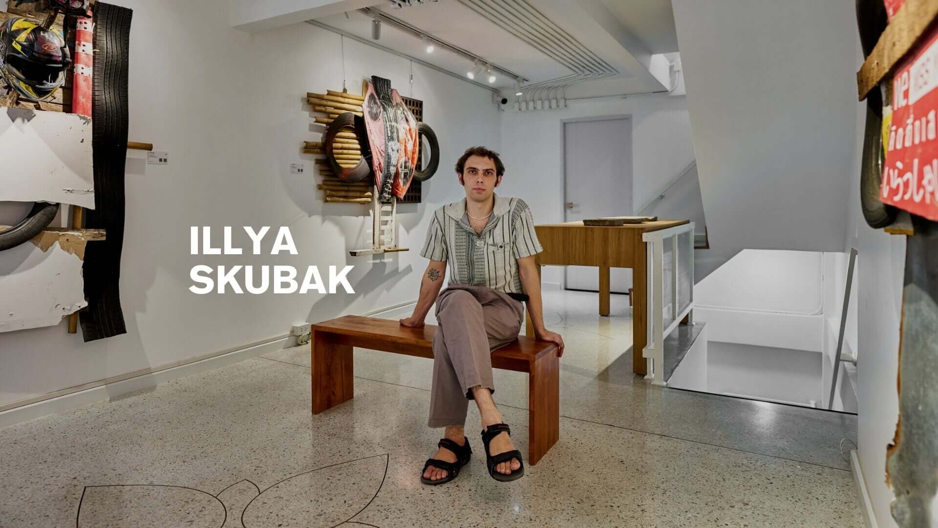 Illya Skubak seated on a wooden bench in an art gallery surrounded by his unique mixed-media artworks.