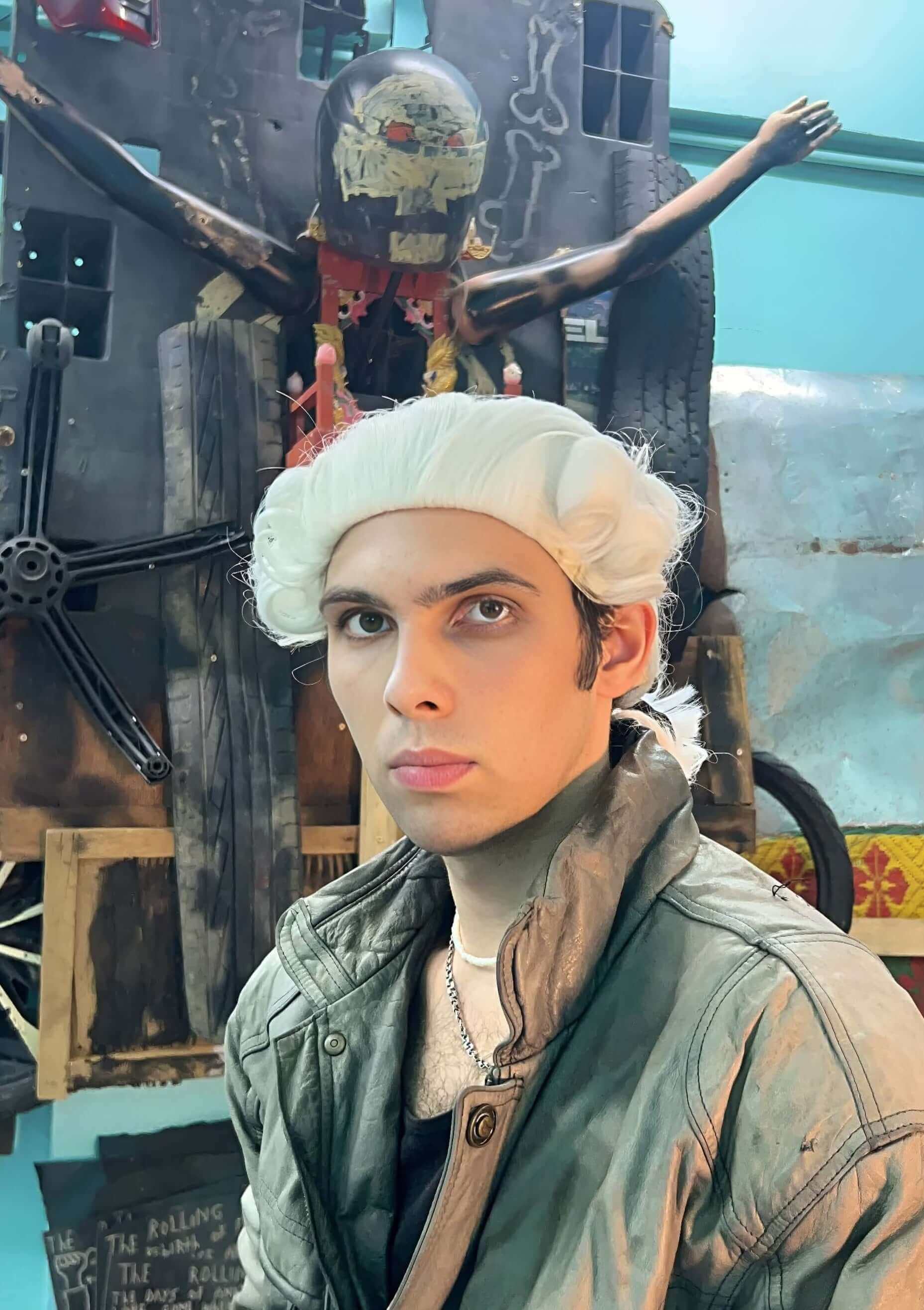 Portrait of Illya Skubak in a white wig and green jacket, set against a colourful artistic backdrop.