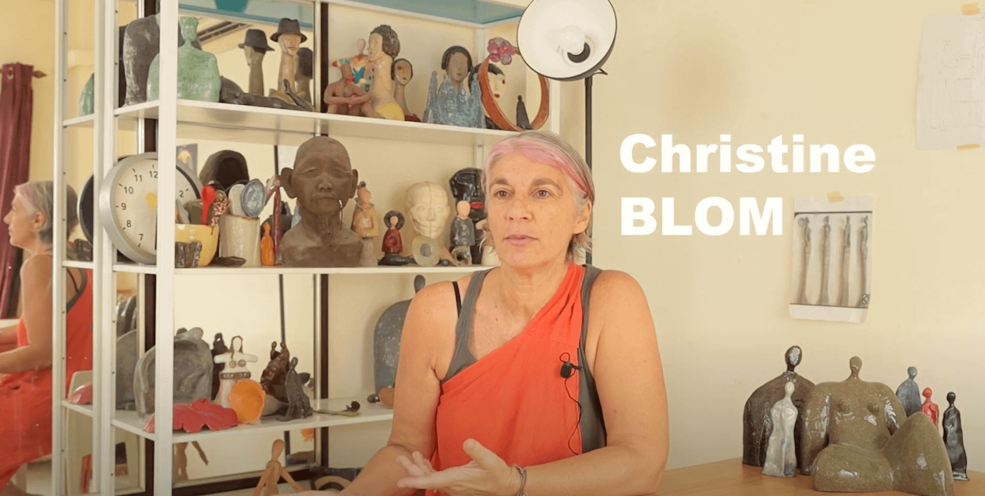 Christine BLOM sitting in her studio with various sculptures displayed on shelves in the background