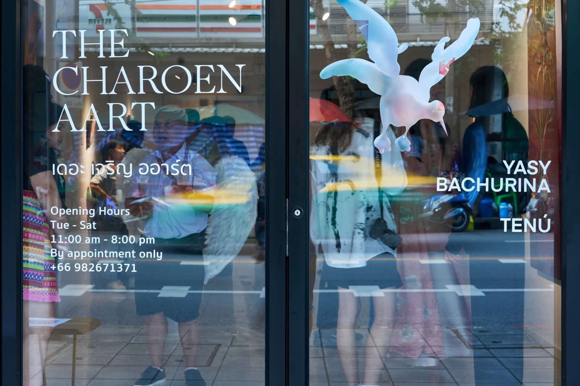 Art gallery entrance displaying exhibition artwork with gallery hours and contact information reflected on the glass door.