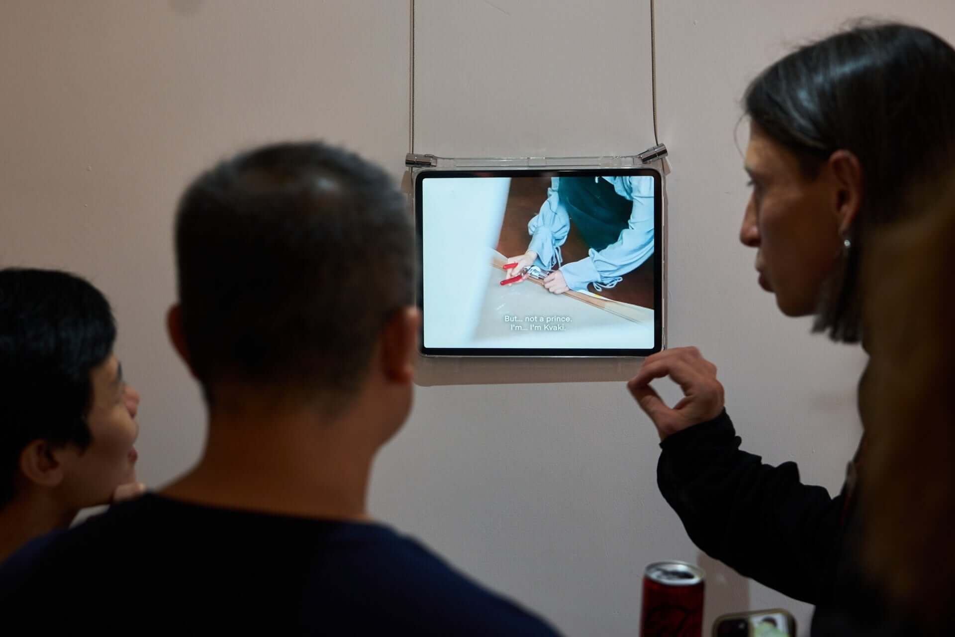 People observing an art installation on a wall-mounted screen displaying hands performing an action.