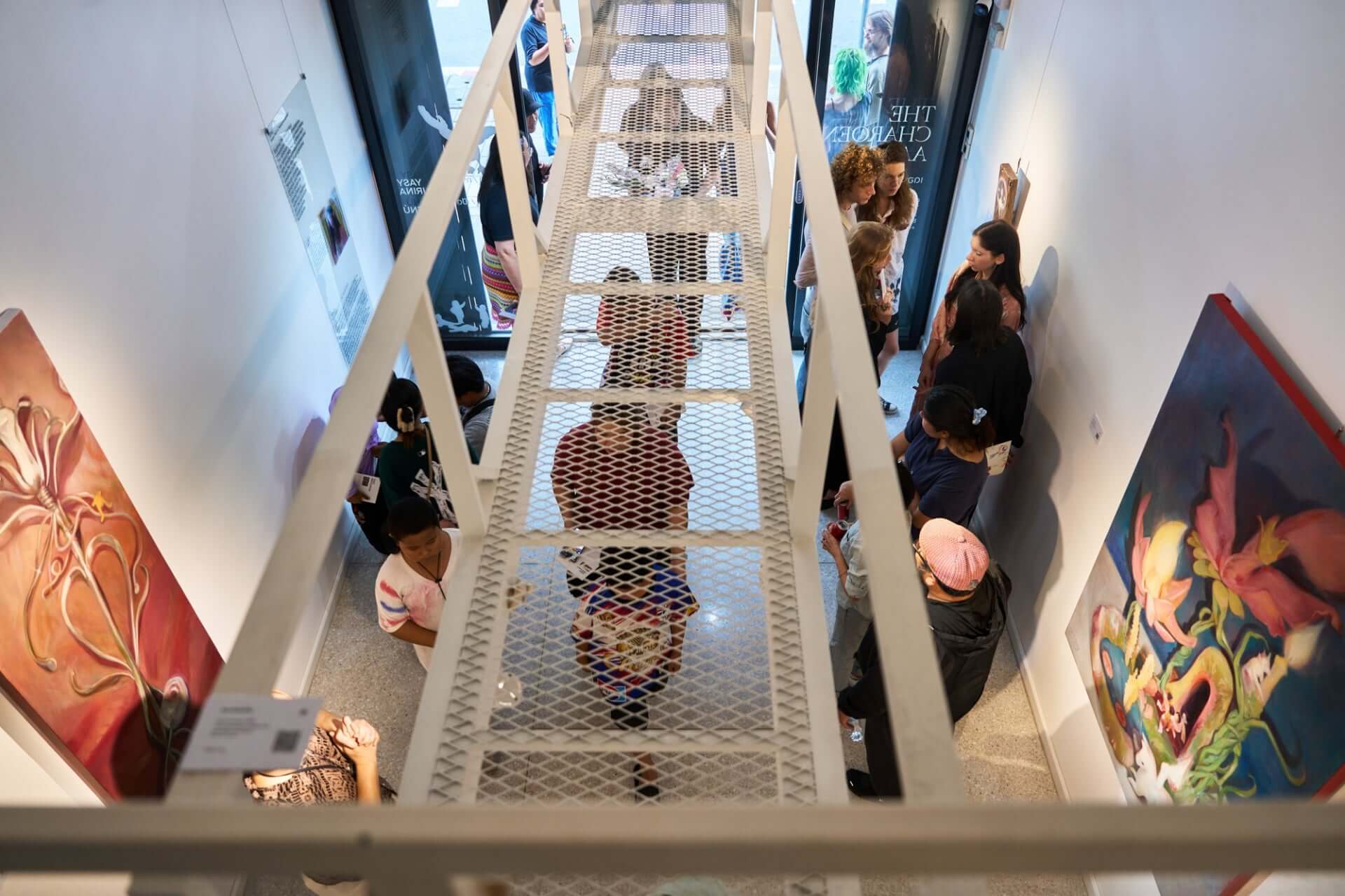 Visitors explore a contemporary art gallery with vibrant paintings, viewed from above through a white metal staircase.