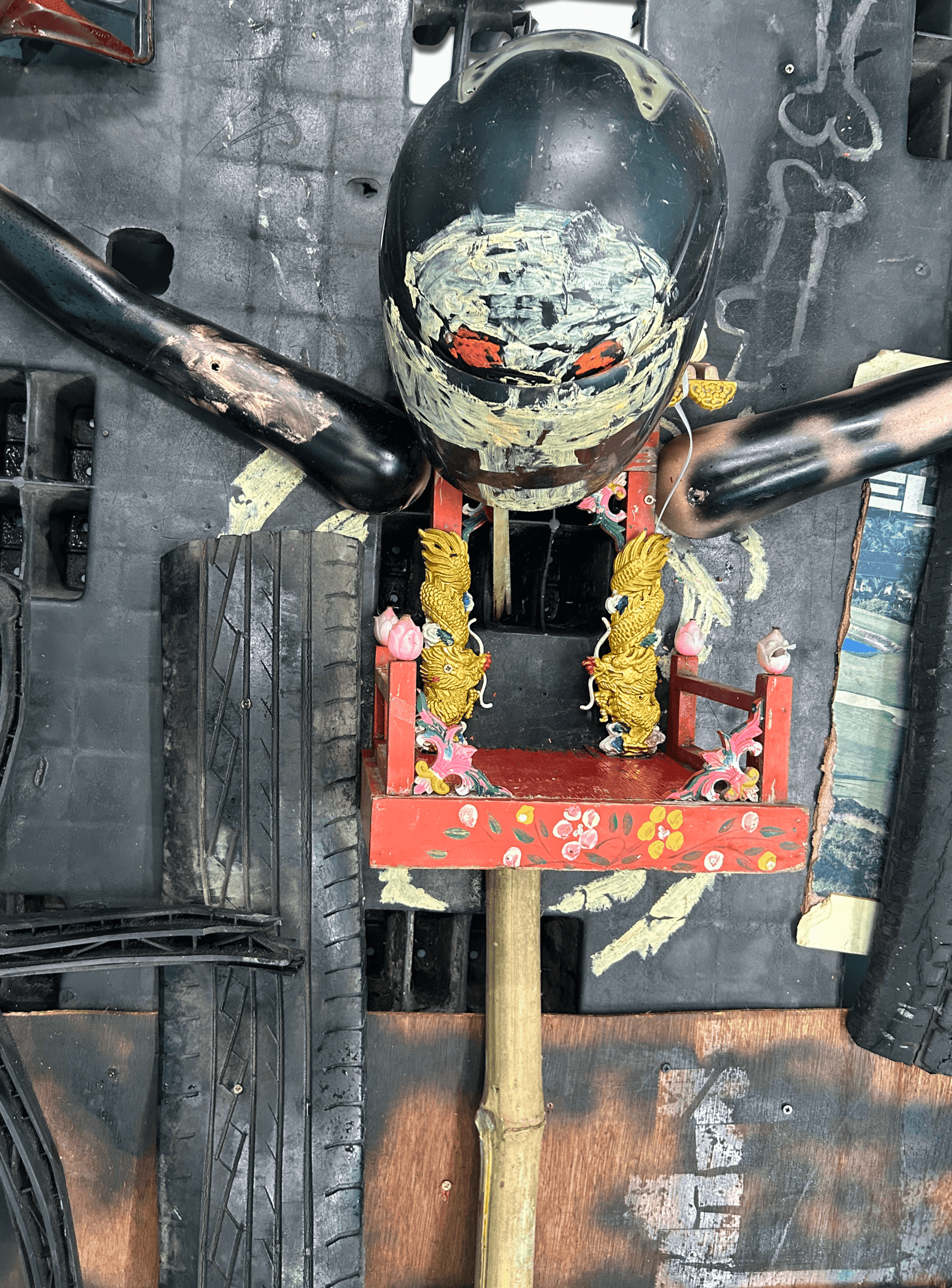 Artistic creation featuring a mask on a wooden structure adorned with colorful decorations and surrounded by tires.