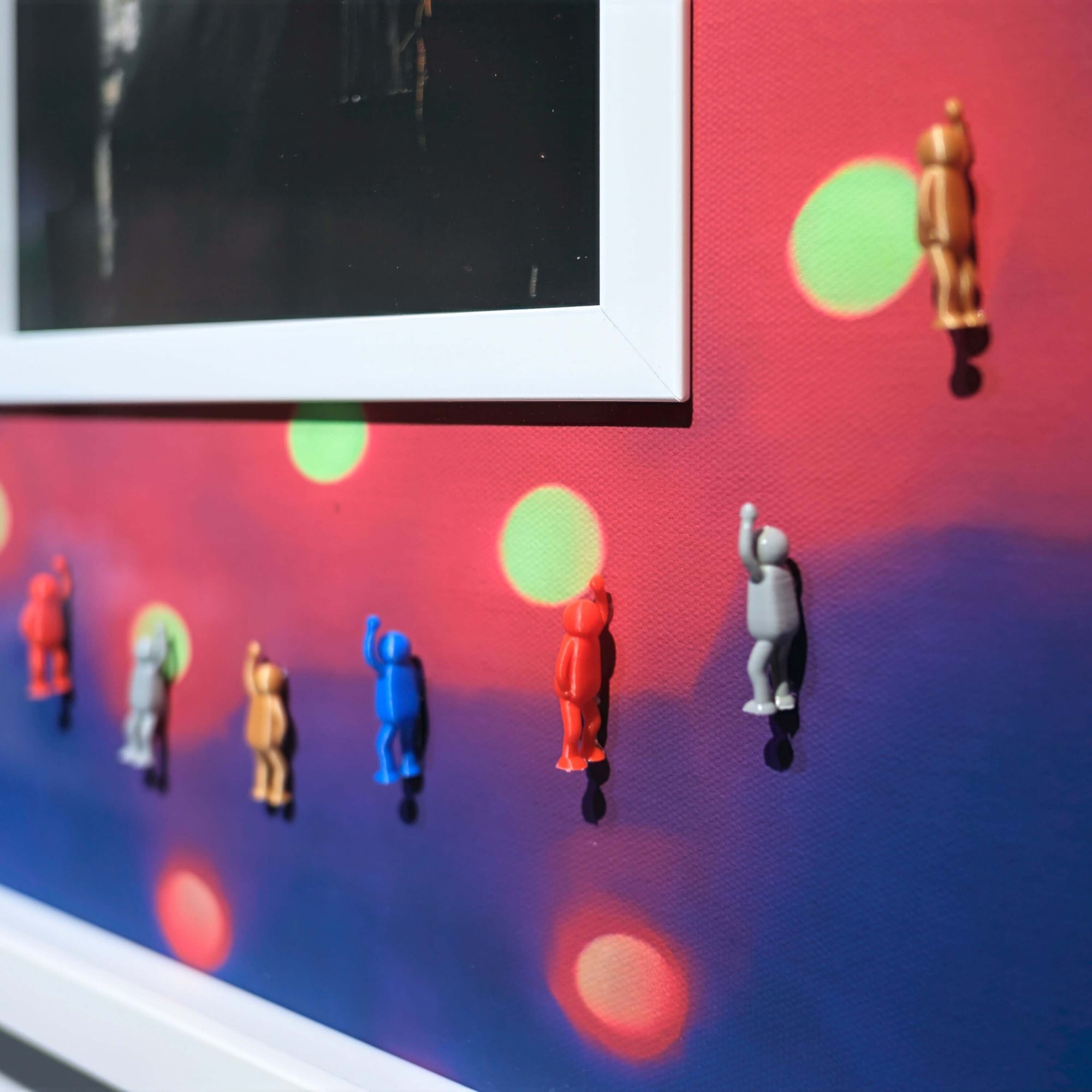 Colorful figurines climbing on a vibrant wall with abstract dot patterns, featuring a framed photo above.