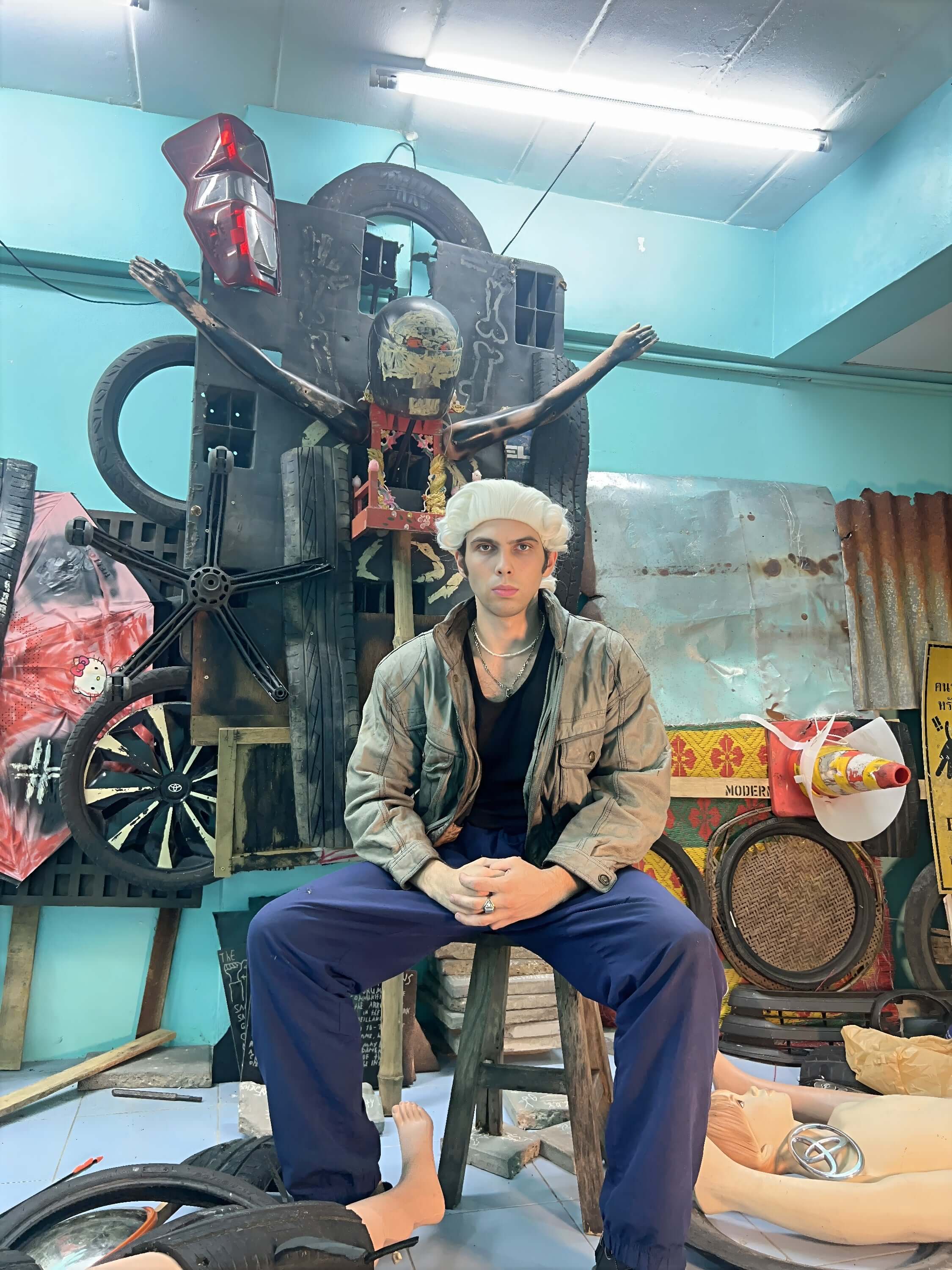 Person in a hat and jacket seated on a stool surrounded by eclectic artwork and props in a creative space.