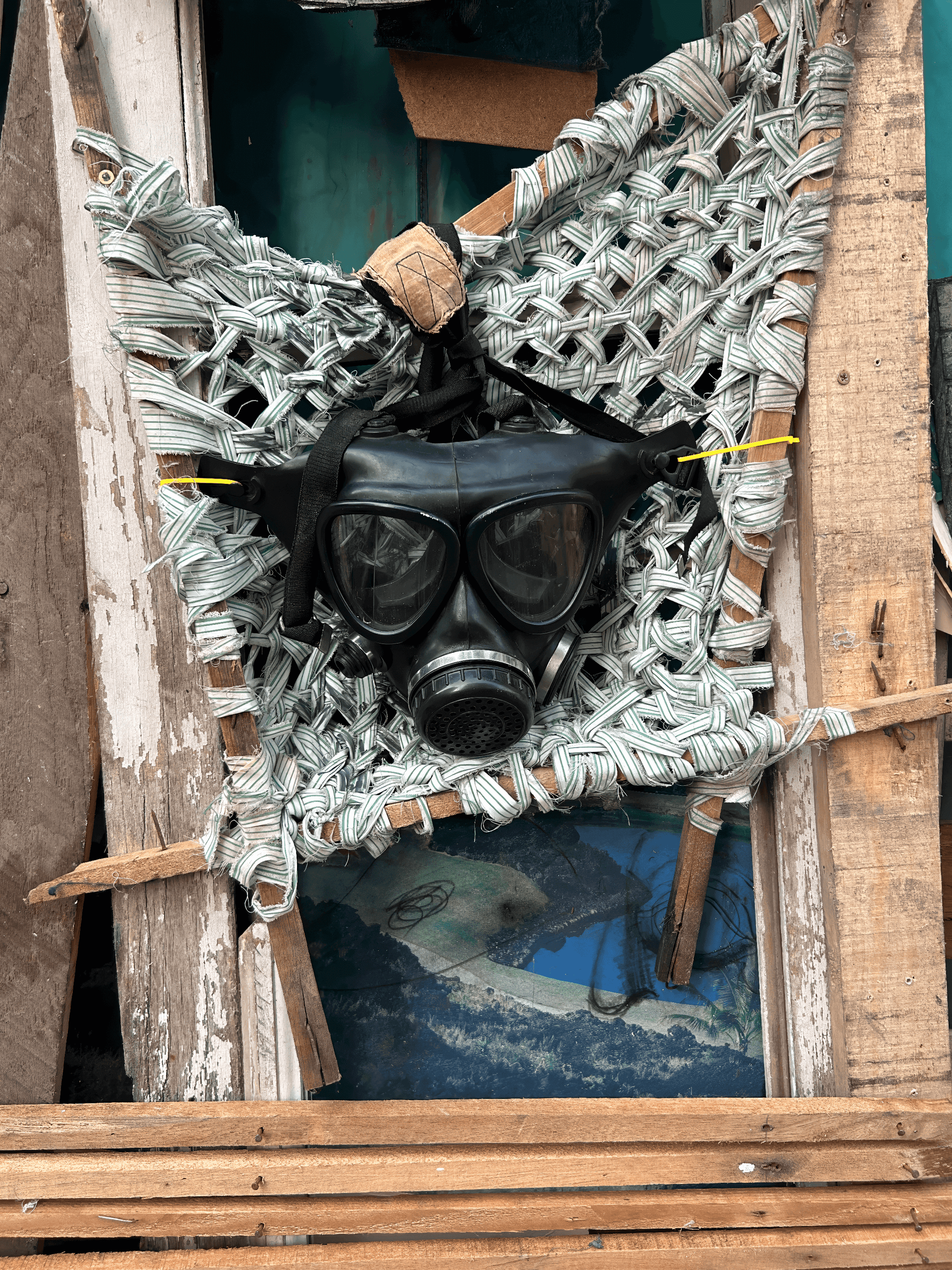 A gas mask displayed in a woven frame made of fabric strips and wooden boards.